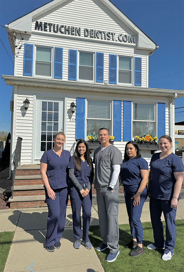 The image shows a group of five individuals standing in front of a building with a sign that reads Metuchen Dental Clinic. They are all dressed in professional attire, suggesting they might be healthcare professionals.