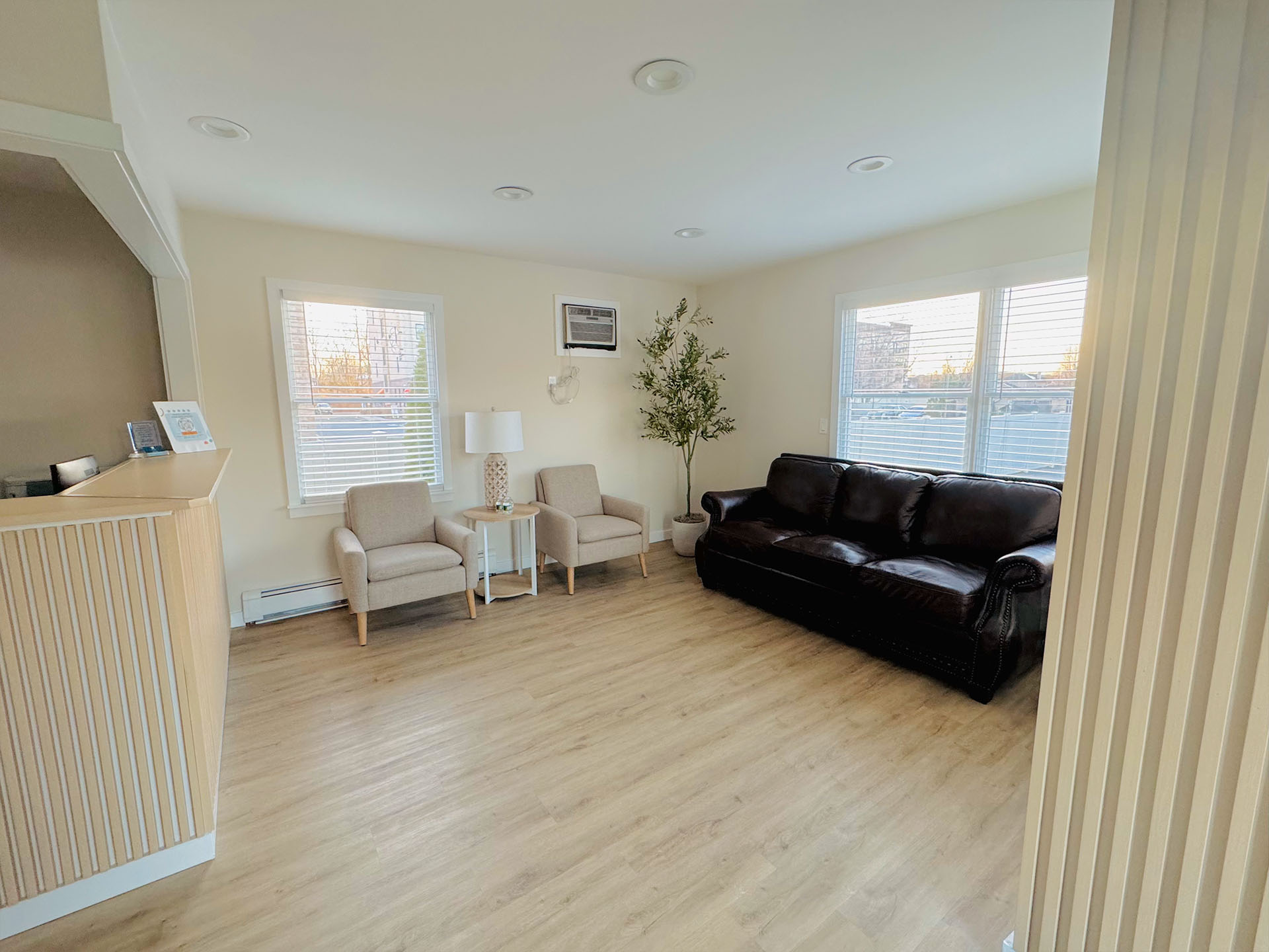 An interior view of a spacious living room with light-colored walls, dark wood flooring, white blinds on windows, a large couch, two chairs, a coffee table, a television, and a dining table with four chairs visible in the background.