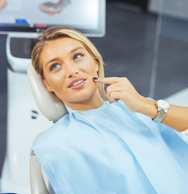 A woman sitting in a dental chair with a smile, looking at the camera while wearing a blue face mask.