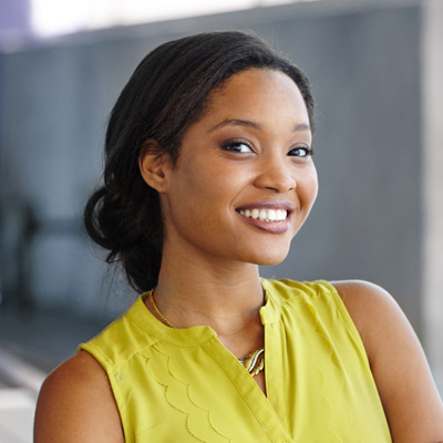 The image depicts a smiling woman with dark hair wearing a yellow top, standing against a blurred background.