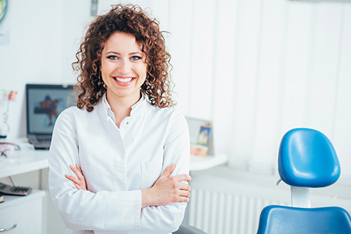The image shows a woman standing in an office environment, smiling at the camera. She has curly hair, is wearing a white blouse, and stands confidently with her hands clasped together.